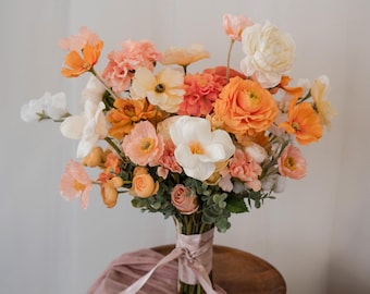May include: A vibrant bouquet of assorted flowers in shades of orange, peach, and white. The arrangement includes ranunculus, poppies, and other blooms, tied with a pink ribbon. The bouquet rests on a wooden surface.