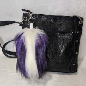Op de afbeelding: Een zwarte crossbody tas van imitatieleer met een sleutelhanger van paarse en witte nep bont bevestigd aan de rits.
