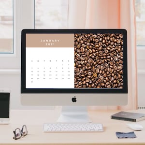 May include: A desktop computer shows a January 2021 calendar and a close-up of coffee beans. The computer sits on a light desk with a smartphone, a coffee mug, and other office supplies. A window and a pink curtain are in the background.