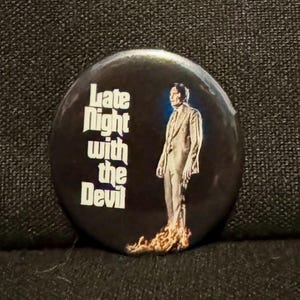 May include: A black and white button with a man in a suit standing in flames. The text "Late Night with the Devil" is printed on the button.