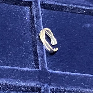 May include: A gold-toned metal ring featuring an open, bypass-style design with two overlapping, slender bands that create a continuous, fluid shape. The ring has a polished, reflective surface and is positioned upright, showcasing its open-ended structure.