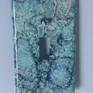 May include: A rectangular light switch wall plate featuring a marbled pattern in shades of blue, white, and dark navy. The cover has a central vertical rectangular cutout for a toggle switch and two small circular holes for mounting screws.