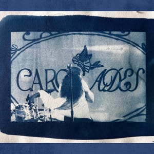 May include: A cyanotype print featuring a performance on stage. The word "CAROADES" is visible in stylized lettering above the performer. The image has a dark blue border and a textured background.