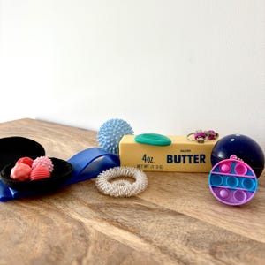 Puede incluir: Una colección de artículos para aliviar el estrés sobre una superficie de madera. Incluye una bola azul con púas, un anillo, una cinta azul, un cuenco negro con formas rosas y rojas, una barra de mantequilla, un llavero, una bola azul y un juguete pop-it rosa y azul.