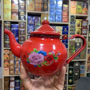 May include: A bright red enamel teapot featuring a floral decal on the front. The teapot has a rounded body, a curved spout, a matching lid with a small knob, and a side handle. The floral design displays pink, red, and blue roses with green leaves.
