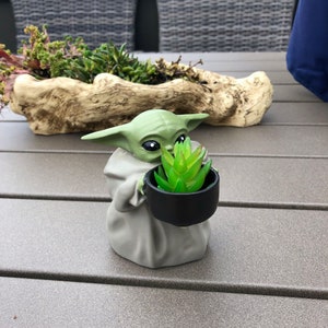 Puede incluir: Una figura verde de Baby Yoda sosteniendo una pequeña maceta negra con una planta suculenta verde. La figura lleva una túnica gris. En el fondo, una gran jardinera decorativa con varias plantas verdes.