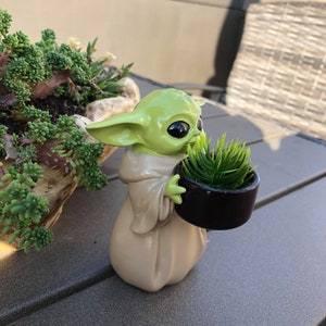 以下が含まれることがあります： 緑色のフィギュアで、小さな黒い鉢植えの植物を持っています。フィギュアは大きな黒い目と尖った耳をしています。キャラクターはベージュのローブを着ています。緑の植物が入った大きなプランターが背景にあります。