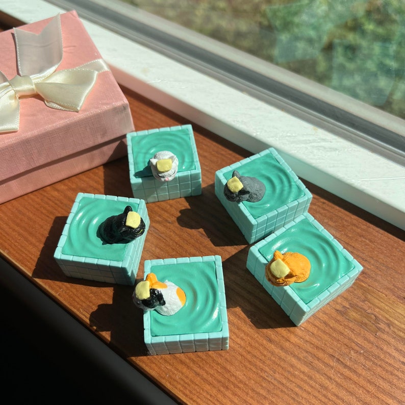 May include: Five miniature ceramic cats wearing yellow hats are floating in turquoise blue pools. Each pool is a square tile with a raised rim. The tiles are arranged in a circle on a wooden surface.