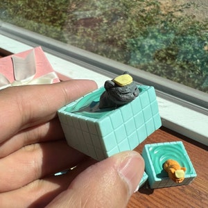 May include: Two small, teal blue, square-shaped bath tubs with a cat figurine in each. The cat in the front tub is orange and the cat in the back tub is gray. Both cats are wearing yellow hats.