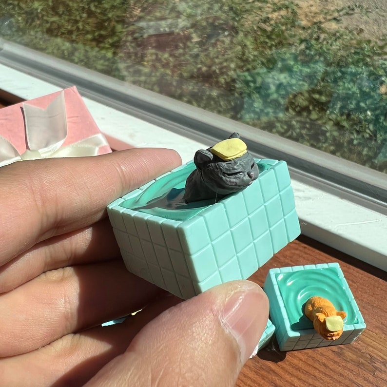 May include: Two small, teal blue, square-shaped bath tubs with a cat figurine in each. The cat in the front tub is orange and the cat in the back tub is gray. Both cats are wearing yellow hats.