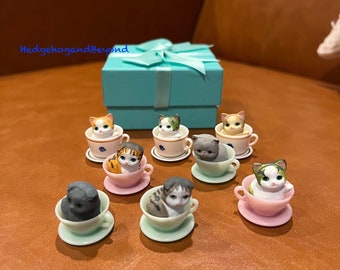 以下が含まれることがあります： ティーカップに座っている9匹のミニチュアセラミック猫のセット。猫は、白、グレー、茶色、黒など、さまざまなポーズと色をしています。ティーカップは花柄で飾られており、ピンク、ブルー、ホワイトの色合いです。