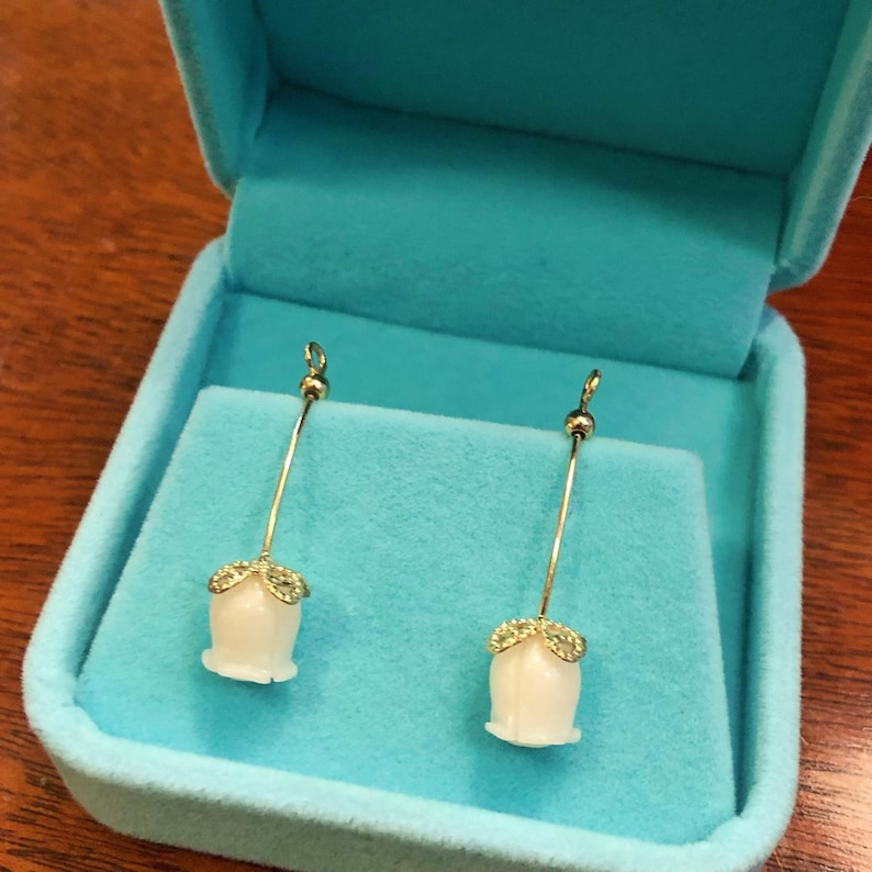 Puede incluir: Un par de pendientes de oro con colgantes blancos en forma de flor que cuelgan de un fino alambre de oro. Los pendientes se muestran en una caja de joyer&iacute;a de terciopelo azul.