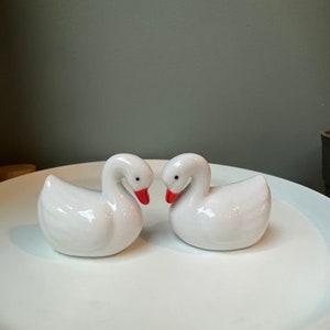 May include: Two white ceramic swan figurines facing each other. The swans have red beaks and are positioned on a white plate.