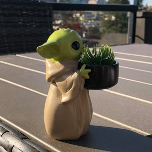 以下が含まれることがあります： 緑とベージュのキャラクターの置物。小さな黒い鉢に緑の植物が入っています。置物は暗い表面にあり、背景にはぼやけた建物と木々があります。