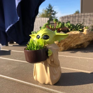 以下が含まれることがあります： 人気のSFフランチャイズのキャラクターの置物で、小さな茶色の鉢に緑の草が入っています。置物は明るい緑とタン色で、大きな黒い目と大きな耳があります。鉢は丸く、濃い茶色です。