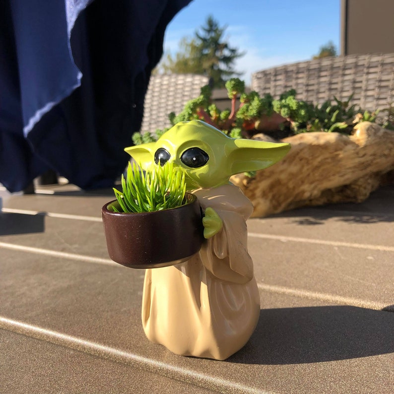 以下が含まれることがあります： 人気のSFフランチャイズのキャラクターの置物で、小さな茶色の鉢に緑の草が入っています。置物は明るい緑とタン色で、大きな黒い目と大きな耳があります。鉢は丸く、濃い茶色です。