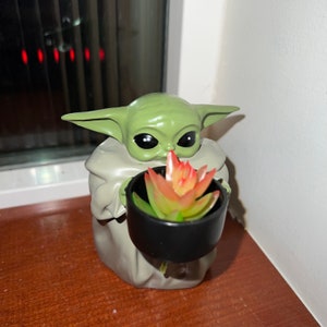May include: A green figurine of a character holding a black pot with a succulent plant. The figurine has large black eyes and pointed ears. The plant has red and green leaves.