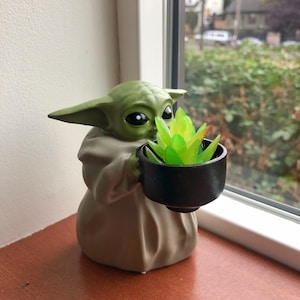 May include: A green figurine of a character holding a small black pot with a green and yellow plant. The figurine is on a brown surface near a window with a view of trees.
