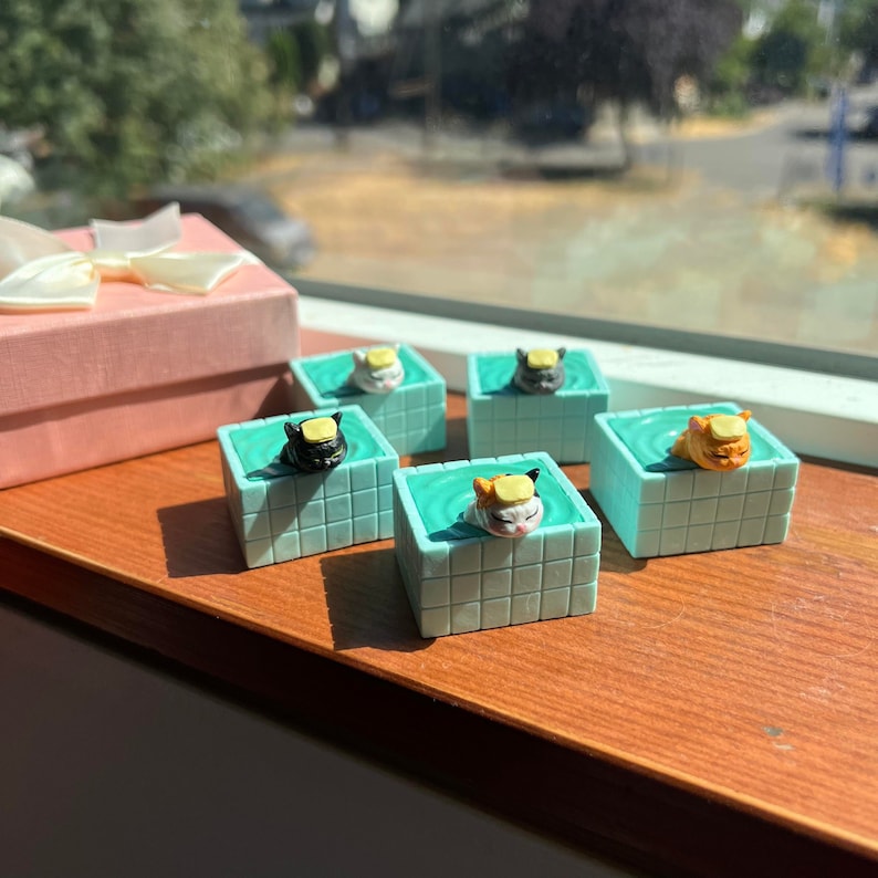 May include: Five miniature ceramic cat figurines in a swimming pool setting. Each cat is wearing a yellow life preserver and is submerged in a turquoise blue pool. The pools are made of a light blue ceramic with a grid pattern. The figurines are sitting on a wooden surface.