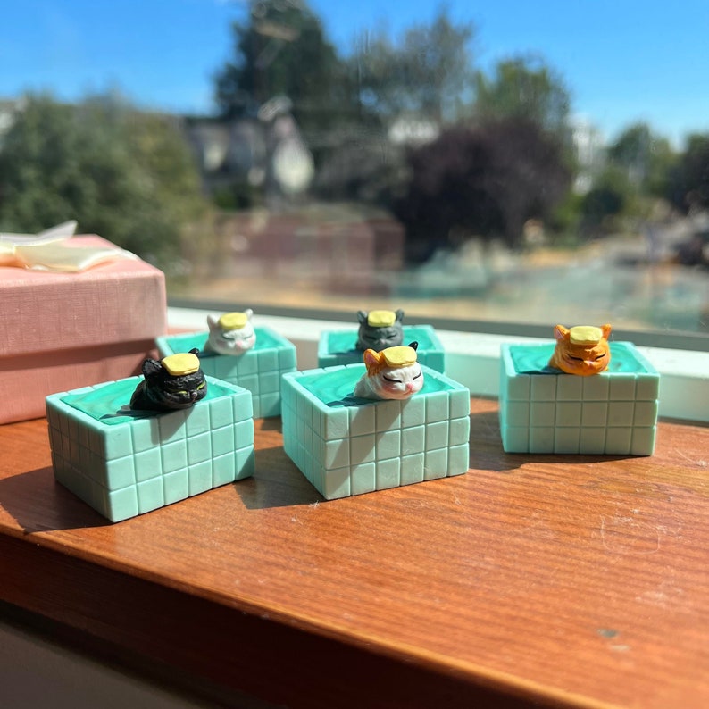 May include: Four miniature ceramic cats wearing hats are sitting in turquoise blue tiled pools. The cats are black, white, orange, and gray. The pools are all the same size and shape.