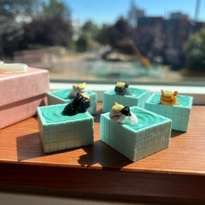 May include: Five miniature ceramic cats wearing yellow hats are floating in turquoise blue pools. Each pool is a square with a grid pattern. The cats are in various poses, some are sleeping, some are swimming. The pools are on a wooden surface.