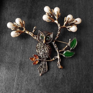 May include: A gold-tone owl brooch with green eyes and rhinestones, perched on a branch with white pearl accents and green leaves.