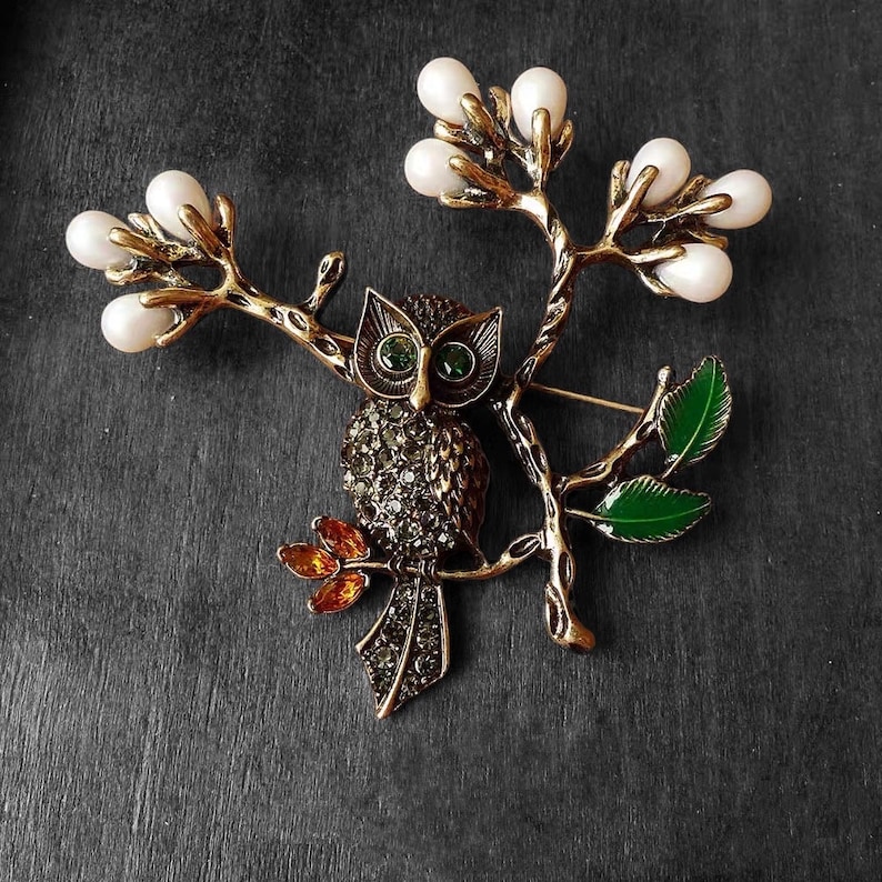 May include: A gold-tone owl brooch with green eyes and rhinestones, perched on a branch with white pearl accents and green leaves.