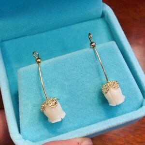 Puede incluir: Un par de pendientes de oro con colgantes en forma de flor blanca. Los pendientes cuelgan de una caja de terciopelo azul.