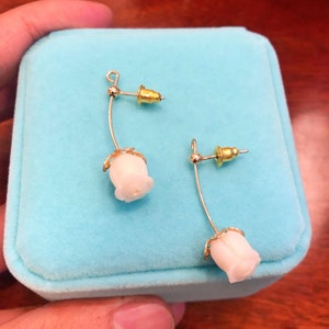 Puede incluir: Un par de pendientes de bot&oacute;n de rosa blanca con detalles dorados y postes plateados. Los pendientes est&aacute;n sobre un fondo de terciopelo azul.