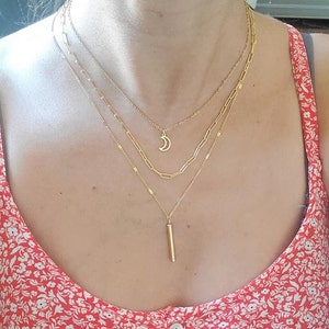 Könnte beinhalten: Ein mehrlagiger Goldketten-Set mit einem Halbmond-Anhänger und einem langen, dünnen Stab-Anhänger.