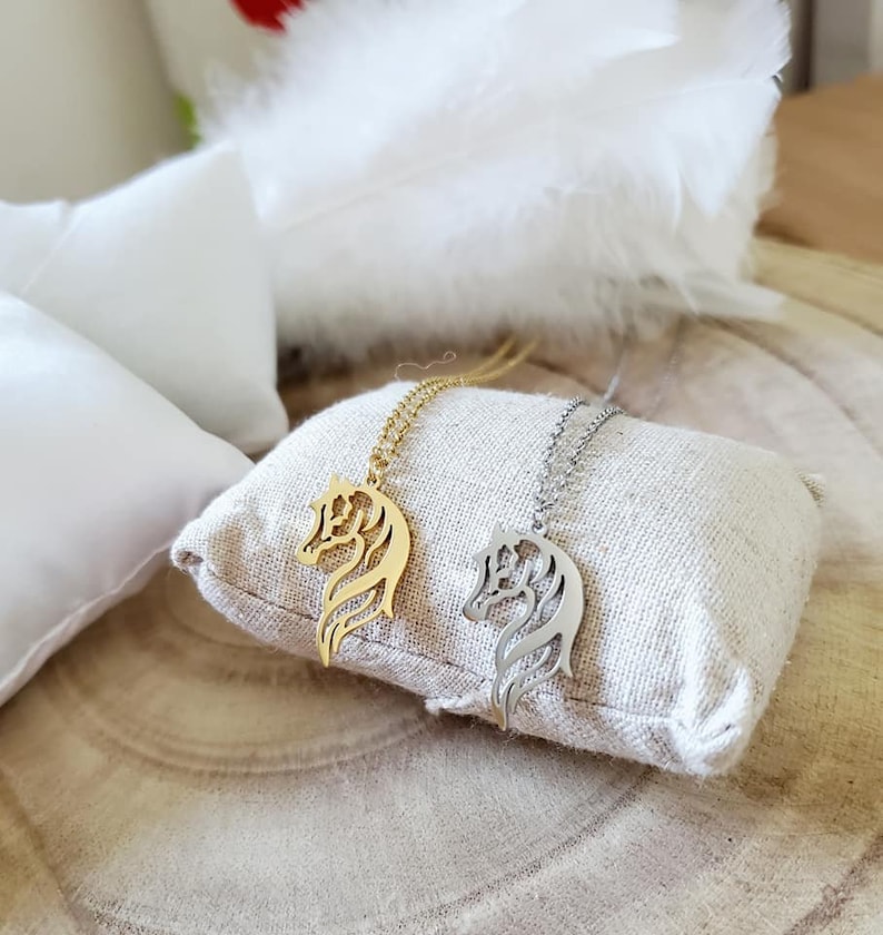 May include: Two horse head necklaces, one gold and one silver, on a beige fabric surface.