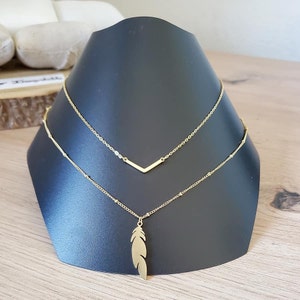 Könnte beinhalten: Eine goldene mehrreihige Halskette mit einem Federnanhänger und einem kleinen V-förmigen Anhänger.