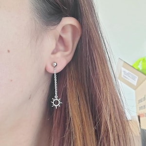 Può includere: Orecchini pendenti in argento con un design a forma di sole e una catena. Gli orecchini sono indossati nell'orecchio di una persona.