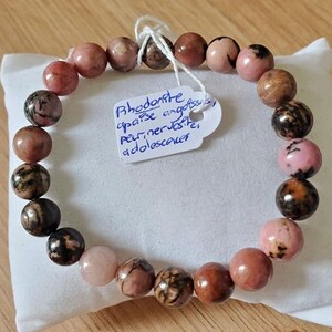May include: A rhodonite beaded bracelet featuring pink, brown, and black spherical beads. A white tag with handwritten text is attached. The bracelet is displayed on a white surface.