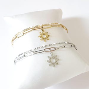 May include: Two gold and silver chain bracelets with sun charm pendants. The bracelets are layered on a white surface.