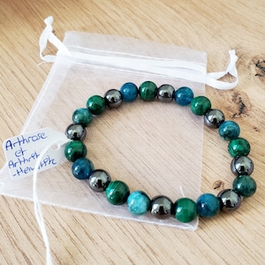 Peut inclure: Un bracelet en perles vertes et noires avec un sac en tissu blanc à cordon. Le sac porte une étiquette manuscrite qui dit "Arthrose et Arthrite -Henriette".