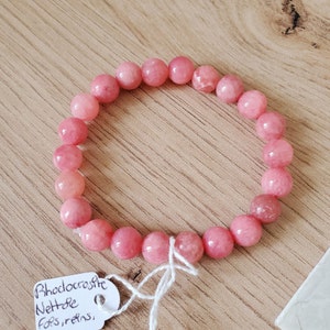 Peut inclure: Un bracelet en rhodochrosite rose avec un cordon blanc noué autour. Le bracelet est accompagné d'une étiquette indiquant "Rhodochrosite Nettle Fossils".