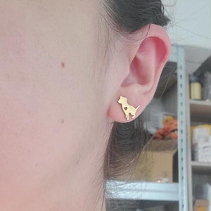Può includere: Orecchini a bottone a forma di cane color oro. Ogni orecchino presenta la silhouette di un cane con un cuore ritagliato. Gli orecchini sono indossati su un orecchio, mostrando le loro dimensioni e il loro design.