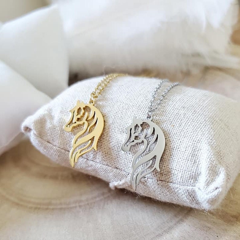May include: Two silver horse head necklaces on a white fabric surface. One necklace is gold and the other is silver.