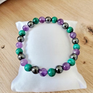 Op de afbeelding: Een armband gemaakt van groene, paarse en zwarte kralen. De kralen zijn rond en hebben een gladde afwerking. De armband ligt op een witte achtergrond.