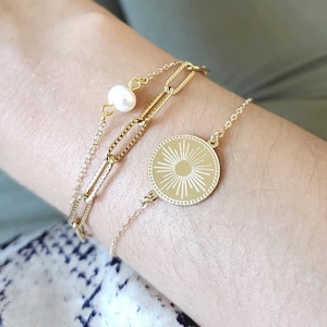 May include: A gold chain bracelet with a pearl charm and a round pendant with a sunburst design.