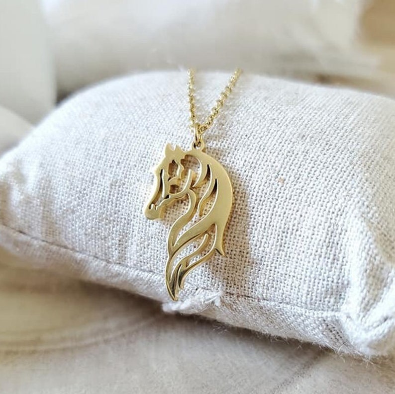 May include: A gold-toned necklace featuring a horse head pendant. The pendant is a cut-out design, with the horse's mane and tail flowing behind it.