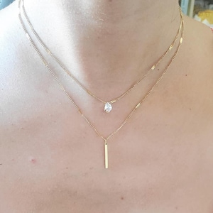 Könnte beinhalten: Eine goldene Kettenkette mit einem tropfenförmigen Anhänger und einem langen, dünnen Stabanhänger.