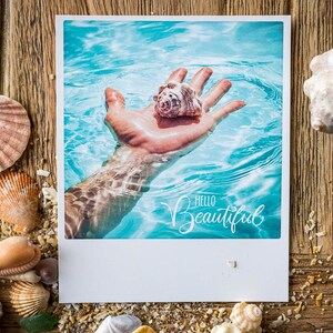 Könnte beinhalten: Ein Polaroid-Foto einer Hand, die eine Muschel in einem blauen Pool hält, mit dem Text "Hello Beautiful" auf dem Wasser.