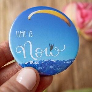 Könnte beinhalten: Ein runder, blauer Button mit dem Schriftzug "TIME IS Now" in weißer Schrift. Der Button zeigt einen Gleitschirmflieger unter einem gelb-orangen Gleitschirmflügel vor blauem Himmel, mit einer Bergkette am unteren Rand.