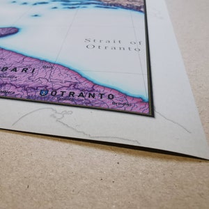May include: A close-up of a map showing the Strait of Otranto, a body of water separating Italy and Albania. The map is printed on paper and framed with a black border.