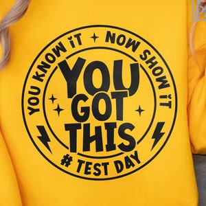 May include: A yellow sweatshirt featuring a black circular design with the text "YOU KNOW IT + NOW SHOW IT, YOU GOT THIS, # TEST DAY". The sweatshirt has long sleeves and a round neckline.