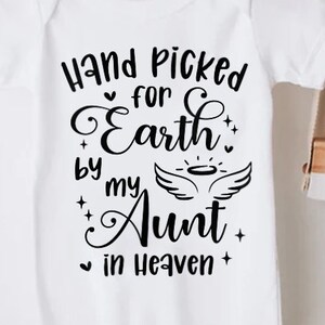 以下が含まれることがあります： 白いベビーロンパースに、黒い文字で「Hand Picked for Earth by my Aunt in Heaven」と書かれています。テキストは小さなハートと星で囲まれています。ロンパースは白い背景に、麦わら帽子と木製のカメラが置かれています。