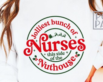 Puede incluir: Sudadera blanca con un diseño circular que dice "Jolliest bunch of Nurses this side of the Nuthouse" en rojo y verde. El diseño incluye acebo y renos. El logotipo de Poetic Panda es visible en la esquina.