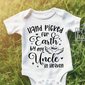May include: White baby onesie with black text that reads "Hand picked for Earth by my Uncle in Heaven". The onesie is lying on green grass.